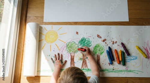 top view of caucasian child drawing green trees and yellow sun with crayons on large paper roll. early education, childhood creativity and art hobby. banner, website header with copyspace.