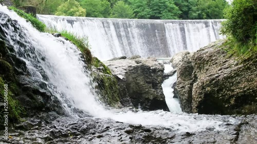 Scenic waterfall flowing over a man-made concrete dam in a lush green forest setting, creating a peaceful and refreshing natural landscape view
