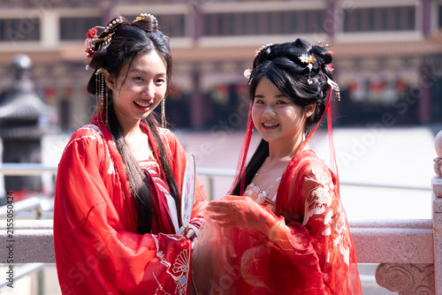 Obraz na płótnie Highlighting traditional Chinese clothing and heritage: two young Asian women in flowing red hanfu gowns, one partly turned away, glancing back smiling, beside red temple courtyard