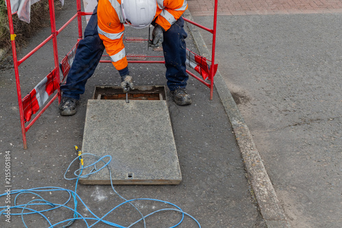 Engineer wearing white hard hat and orange high visibility jacket lifting chamber cover slab back into place  at telecoms works in residential street