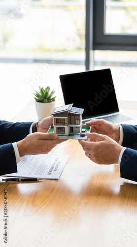 Hands exchanging a model house on a table with a laptop and contract in the background. This scene symbolizes a real estate transaction or business deal, suggesting a property handover.