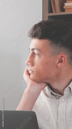 Portrait of a pensive and tired young man looking away from his laptop screen, feeling bored and unmotivated while studying or working on a difficult task in his home office or library