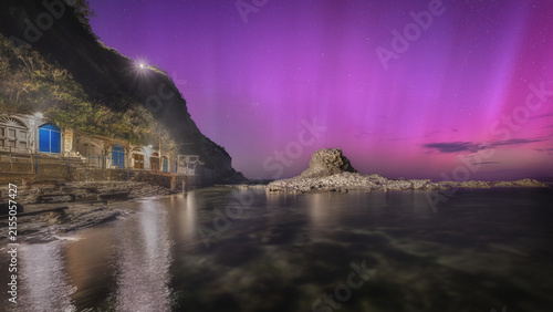 View of the Chair of the Pope rock formation illuminated by a vibrant purple aurora borealis above the rocky coastline and grottos of Ancona, Marche, Italy.