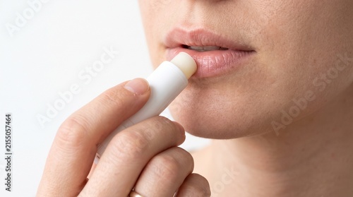 Close-up shot of a woman applying lip balm to her lips. The woman's hand is visible, holding the white tube of lip balm. Her lips are slightly parted, and she is focused on the task