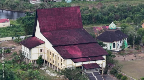 A large traditional Banjar house with a red roof sits amidst trees and grass, south Kalimantan