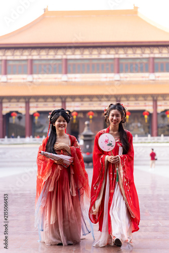 Fotografía Two young Chinese women in matching red hanfu costumes, walking, one glancing sideways at partner, at temple courtyard plaza, highlighting traditional Chinese clothing and heritage