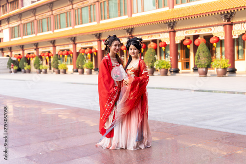 Fotografie Two young Chinese women in red silk hanfu dresses, full body standing, weight shifted, relaxed smile, at red pillared corridor, hanging lanterns, in a traditional hanfu culture photo shoot