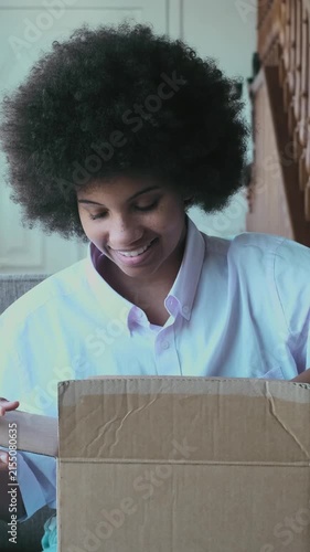 Joyful young woman with an afro hairstyle unboxing a cardboard package after an online purchase, showing curiosity and happiness while opening her long-awaited delivery at home