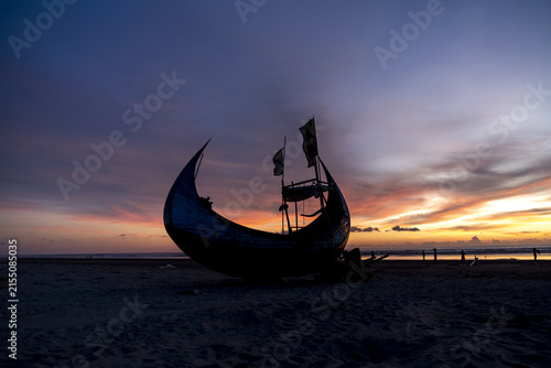 View of a traditional wooden fishing boat silhouetted against a vibrant sunset sky on the sandy shore of Inani Beach, Cox's Bazar District, Chittagong Division, Bangladesh.