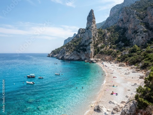 A high-angle view of a pebble beach with turquoise sea, boats anchored in the water, and sunbathers resting on the shore under tall limestone cliffs.