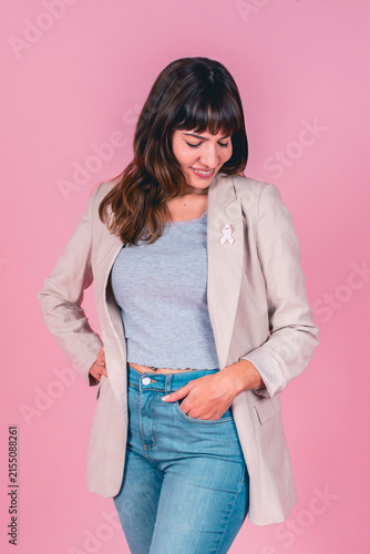 Smiling woman in beige blazer looking down at pink ribbon for breast cancer awareness on pink background