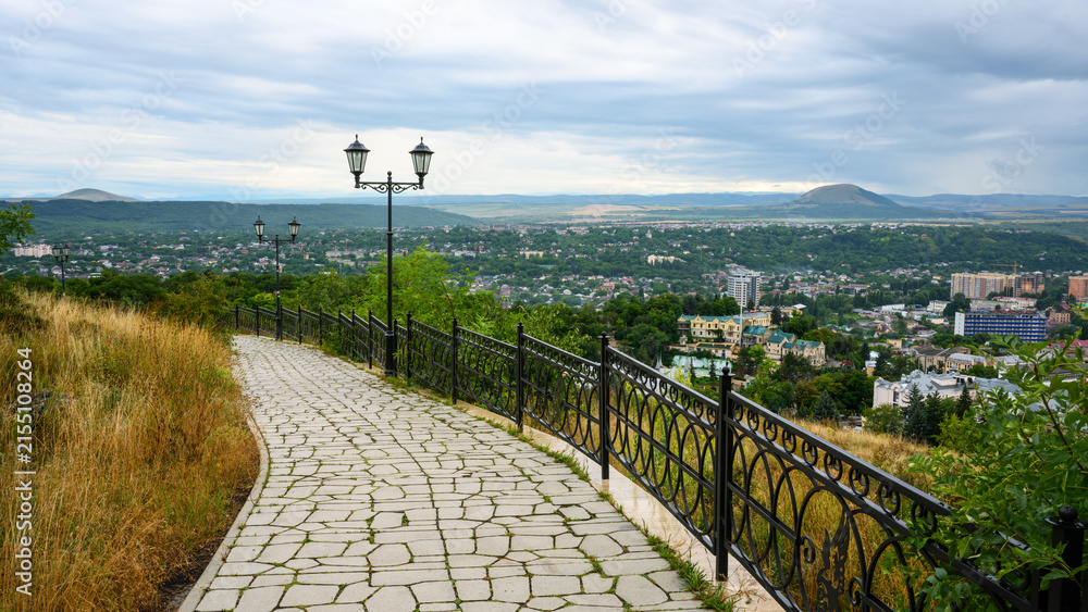 Obraz premium Paved walkway with a metal fence and vintage street lamps overlooking a city of Pyatigorsk, Russia. Panoramic urban landscape in a valley surrounded by mountains. Scenic elevated viewpoint
