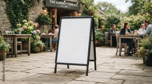 Blank white sandwich board sign standing on a stone patio outside a restaurant with blurred dining customers