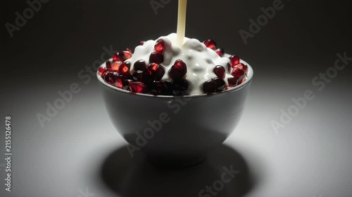 Bright red pomegranate arils inside a white ceramic bowl, thick cream streaming down from above