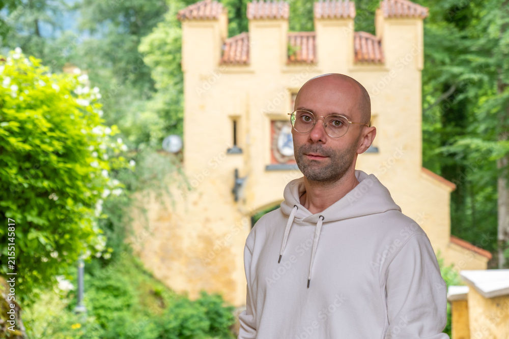 Fototapeta premium young man with round glasses wearing white hoodie, tourist standing beneath medieval castle background, historic heritage landmark, outdoor portrait, contemplative expression, casual streetwear style
