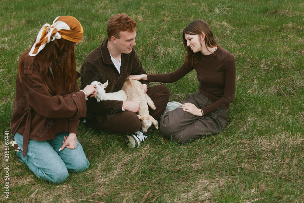 Obraz premium Casual outdoor scene with a family and friends petting a curious goat kid on a sunny grassy field, relaxed smiles, warm tones, gentle breeze, tender moment of countryside lifestyle