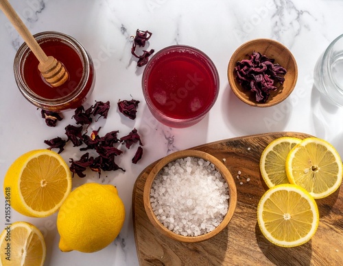 A glass of red roselle drink sits beside lemon, honey, sea salt, and dried roselle on a marble surface in an overhead beverage preparation scene