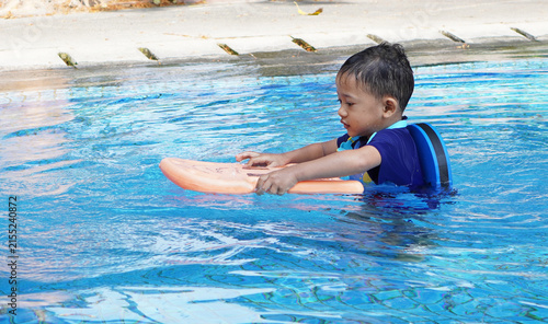 Quadro su tela A young Asian boy is concentrating on learning to swim, wearing a blue back float and holding an orange plastic float in front of him to keep from sinking