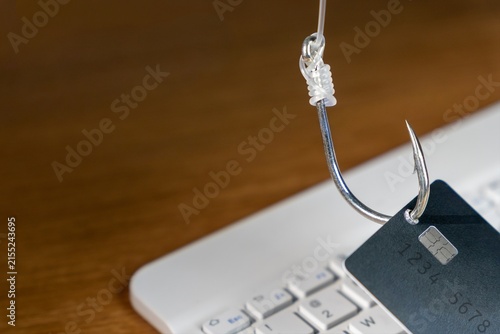 Fishing hook piercing a credit card over a blurred laptop keyboard. Online phishing scams, cybersecurity threats, digital fraud, and internet identity theft concept.
