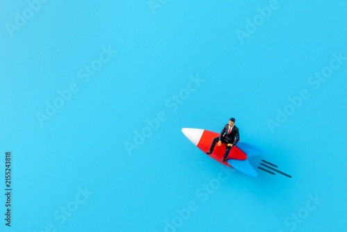 Photography Miniature businessman figurine riding a rocket ship across a blue background
