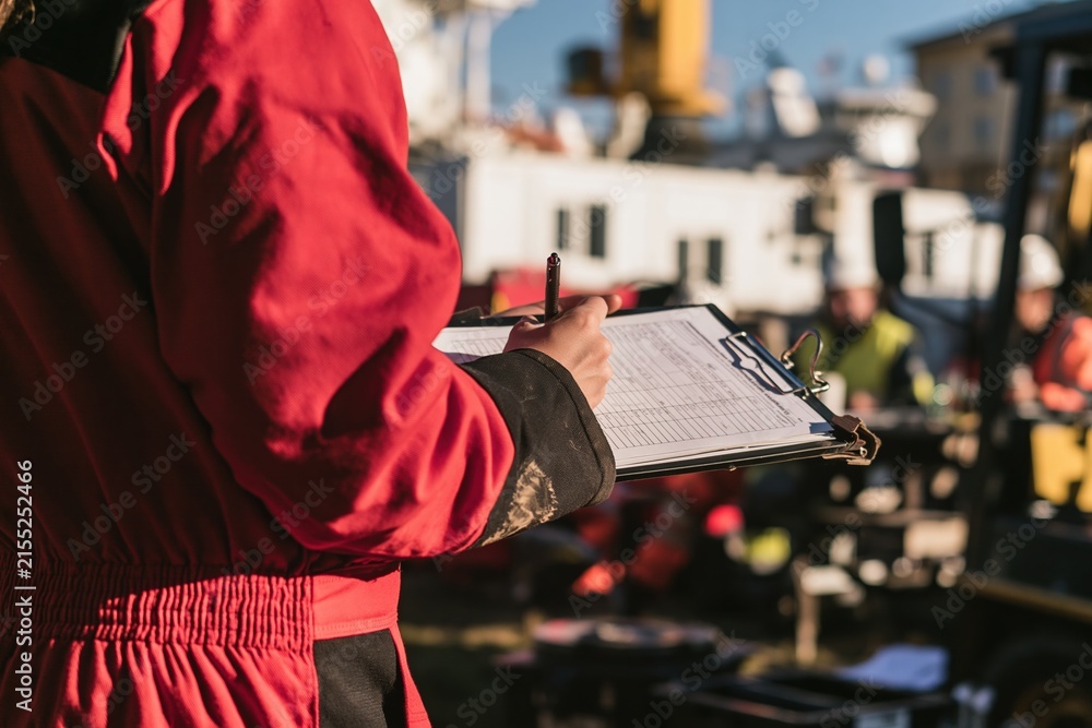Obraz premium Red high-visibility coverall sleeve and hand writing on a clipboard, focused on a weathered risk assessment form for site audit and safety communication