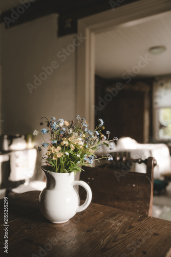 Laane County, Estonia - 25 May 2023: View of a white ceramic pitcher filled with blue and yellow wildflowers on a rustic wooden table in a cozy interior.