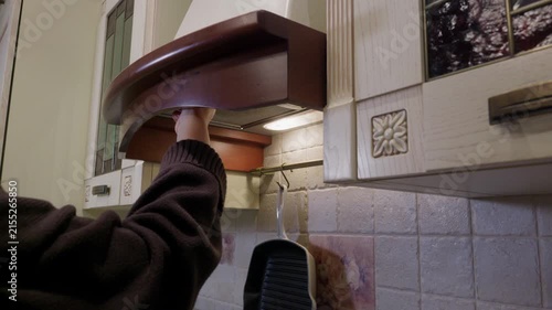 Person in black sweater reaches up to deactivate brown wooden range hood above stovetop in residential kitchen with tiled backsplash.