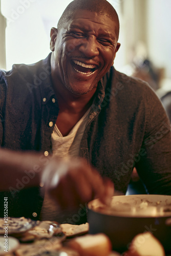 A Black man is laughing wholeheartedly while sitting at a table, about to eat. His eyes are closed, and his head is tilted back as he bursts into laughter.