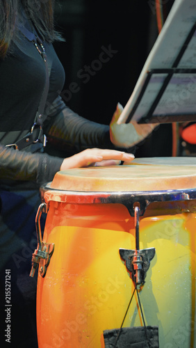 Closeup of musician hands on conga drum under warm stage lighting. Vibrant orange yellow drum contrasts with dark outfit. Music stand visible in background
