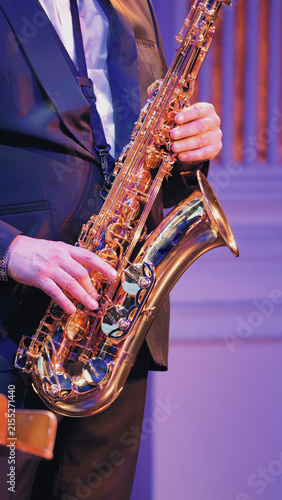 Closeup of musician playing golden saxophone under dramatic purple stage lights. Formal suit, hands on keys, reflective brass surface. Live performance mood, concert atmosphere, music event setting