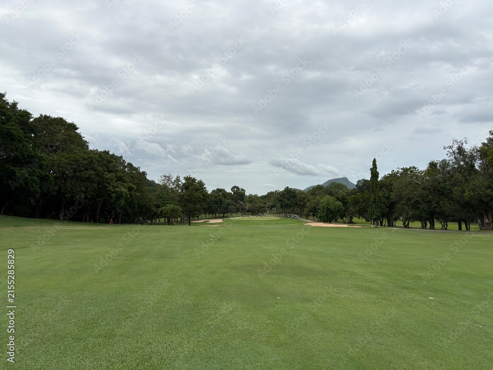 Fototapeta premium Wide golf fairway with sand bunkers and distant mountain under cloudy sky in Thailand