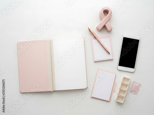 Flat lay composition with blank open notebook pen smartphone memo pad pill organizer and pink ribbon on white background surface.