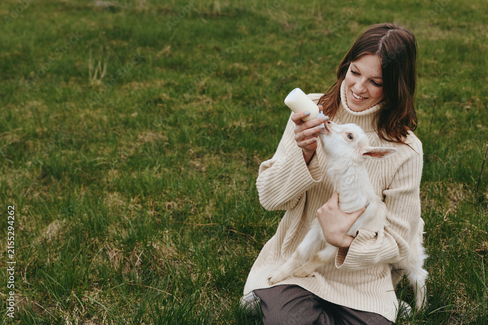 Obraz premium A woman in a cozy knitted sweater feeds a white baby goat with a bottle in a countryside field, capturing a warm lifestyle moment and gentle animal bonding.