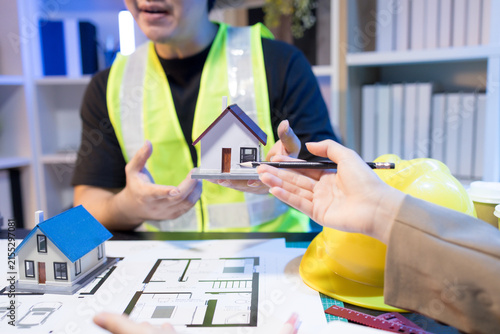 Two people are working together at a dimly lit desk. They are working on a home improvement project, the lighting adding to their focused expressions. Books and notebooks are stacked on shelves behind