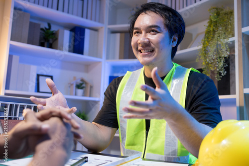 Two people are working together at a dimly lit desk. They are working on a home improvement project, the lighting adding to their focused expressions. Books and notebooks are stacked on shelves behind