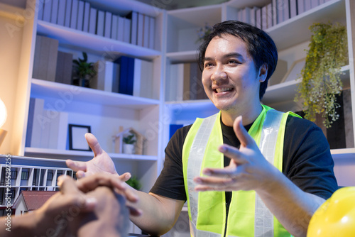 Two people are working together at a dimly lit desk. They are working on a home improvement project, the lighting adding to their focused expressions. Books and notebooks are stacked on shelves behind