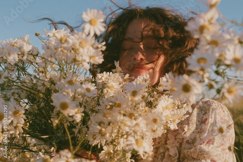 Woman surrounded by white daisies in a sunny meadow, with flowers framing her face and warm light creating a dreamy, natural summer portrait.