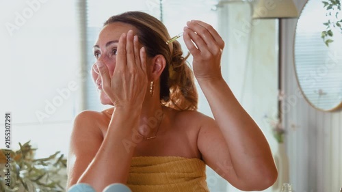 Smiling mature woman applies facial oil with a pipette in the bathroom, enjoying a healthy skincare routine and confident self care at home.