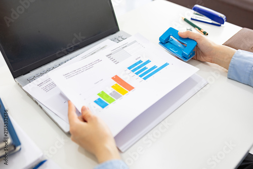 ands hold colorful charts beside a laptop. Fingers press a stapler, securing reports for analysis. Office desk holds a mouse, pen, and notebook. Focused work suggests data review or presentation prep
