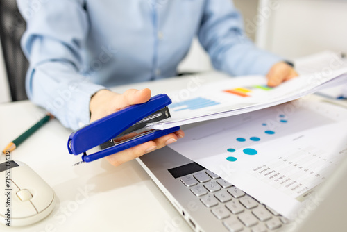 ands hold colorful charts beside a laptop. Fingers press a stapler, securing reports for analysis. Office desk holds a mouse, pen, and notebook. Focused work suggests data review or presentation prep
