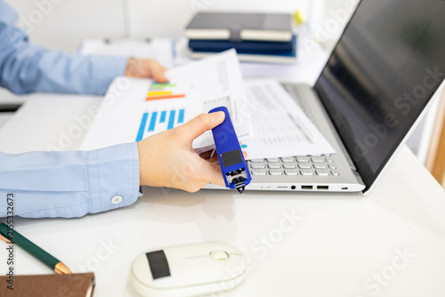 ands hold colorful charts beside a laptop. Fingers press a stapler, securing reports for analysis. Office desk holds a mouse, pen, and notebook. Focused work suggests data review or presentation prep
