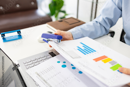 ands hold colorful charts beside a laptop. Fingers press a stapler, securing reports for analysis. Office desk holds a mouse, pen, and notebook. Focused work suggests data review or presentation prep
