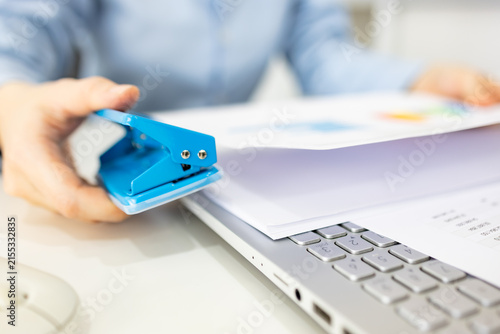 ands hold colorful charts beside a laptop. Fingers press a stapler, securing reports for analysis. Office desk holds a mouse, pen, and notebook. Focused work suggests data review or presentation prep
