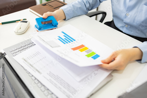 ands hold colorful charts beside a laptop. Fingers press a stapler, securing reports for analysis. Office desk holds a mouse, pen, and notebook. Focused work suggests data review or presentation prep
