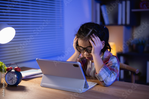 Tired young girl wearing glasses sitting at desk using tablet with frustrated expression late at night under soft lamp light in cozy room setting