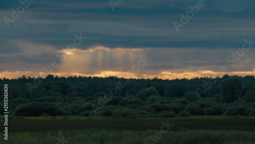 Sunbeams breaking through dark storm clouds above forest and wetland at sunset