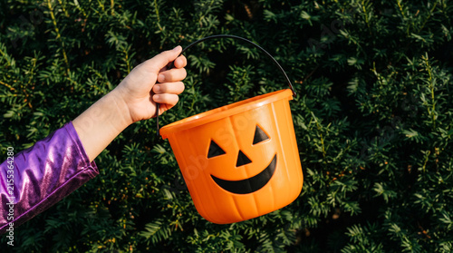 Child hand holding an orange plastic jack-o-lantern candy bucket for Halloween trick or treat