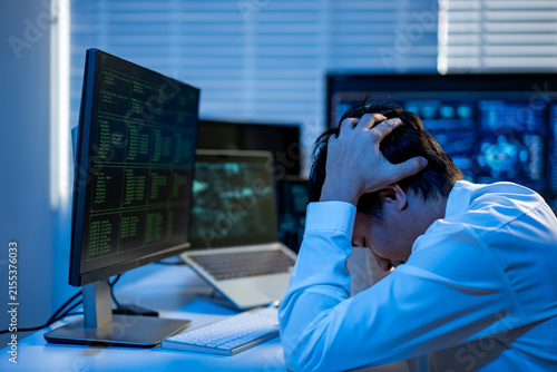 Stressed office worker sitting at a desk with computer screens, holding his head in frustration after long hours of work