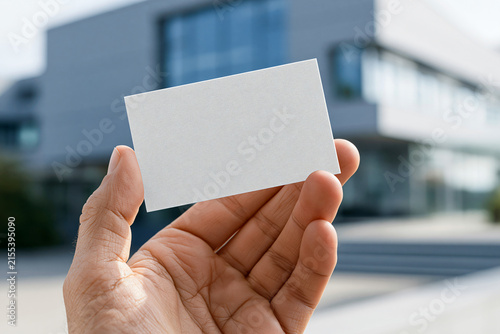 A Hand Holds a Blank Business Card Outside a Modern Building A close-up shot features a person's hand holding a blank white business card in front of a contemporary architectural structure.