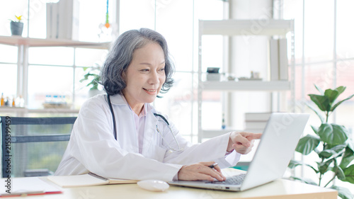 Asian smiling older woman doctor wearing medical coat stethoscope sitting working with laptop computer in healthcare office professional physician services hospital clinic wellness trust patient care.